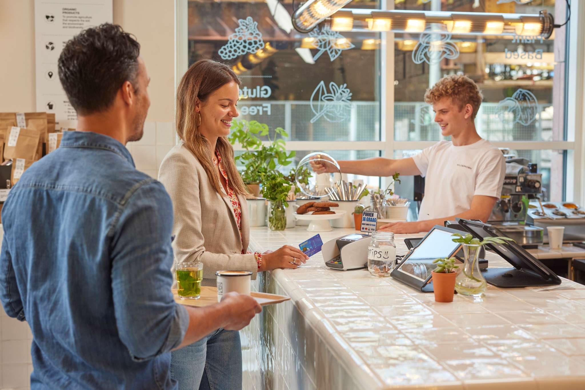 Vrouw rekent glimlachend af met betaalpas aan lichte koffiebar, terwijl barista en wachtende klant ontspannen bij de toonbank staan.