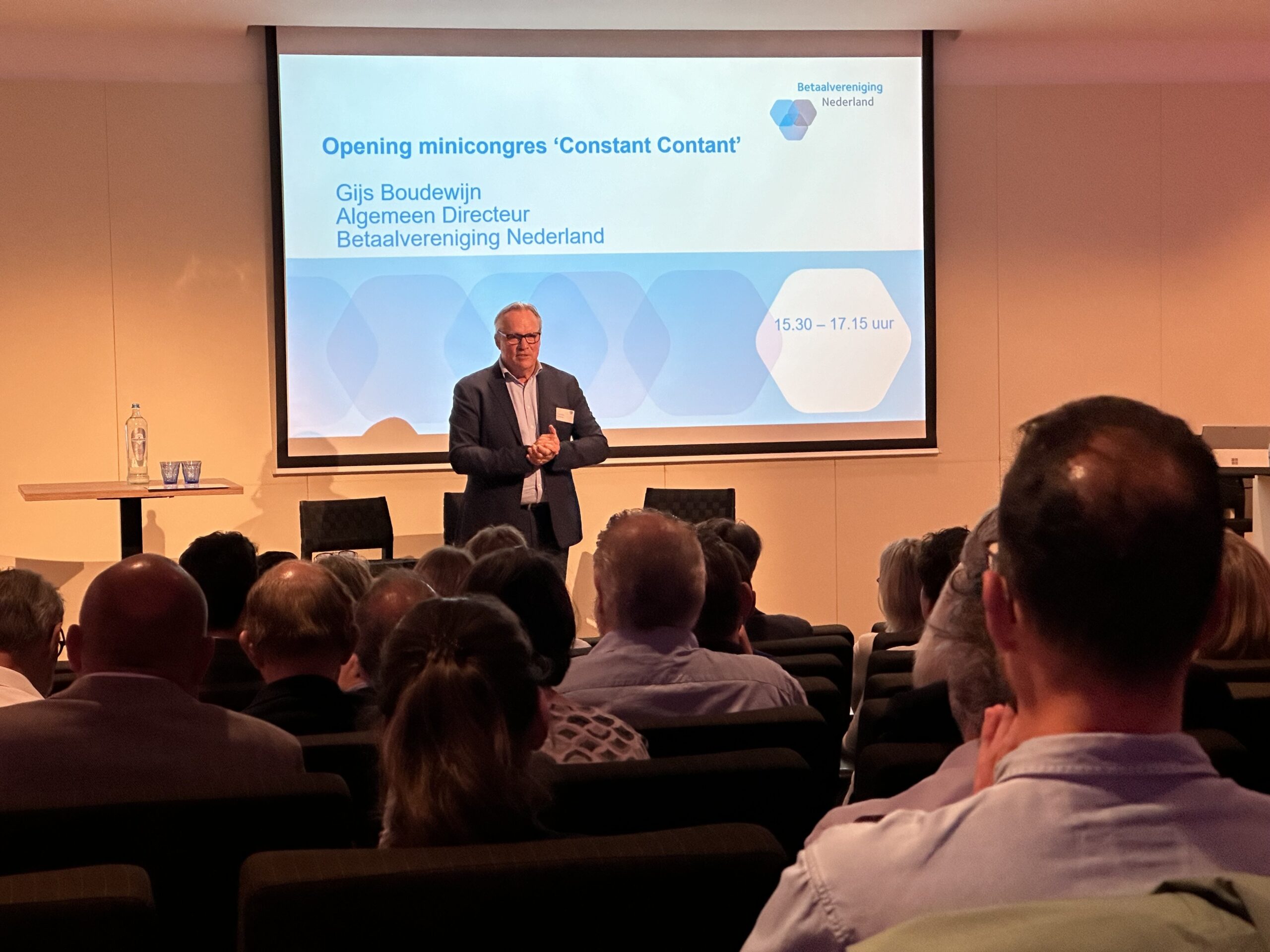 Gijs Boudewijn opent het minicongres ‘Constant Contant’ van Betaalvereniging Nederland; hij staat voor een groot projectiescherm met de titel van de bijeenkomst terwijl het publiek in de zaal op hem gericht zit.