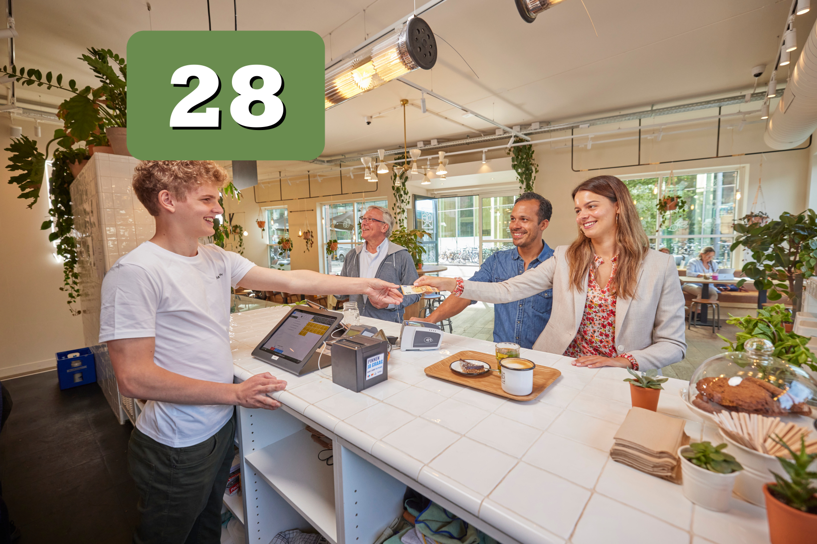 Klant rekent af aan de toonbank van een lichte koffiebar terwijl een medewerker een betaalpas aanneemt, met zichtbaar een grafisch blok met het getal 28.