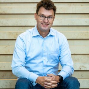 Hans Overeem, Head of Payments bij ING, zit ontspannen met gevouwen handen en kijkt professioneel in de camera tegen een rustige houten achtergrond.