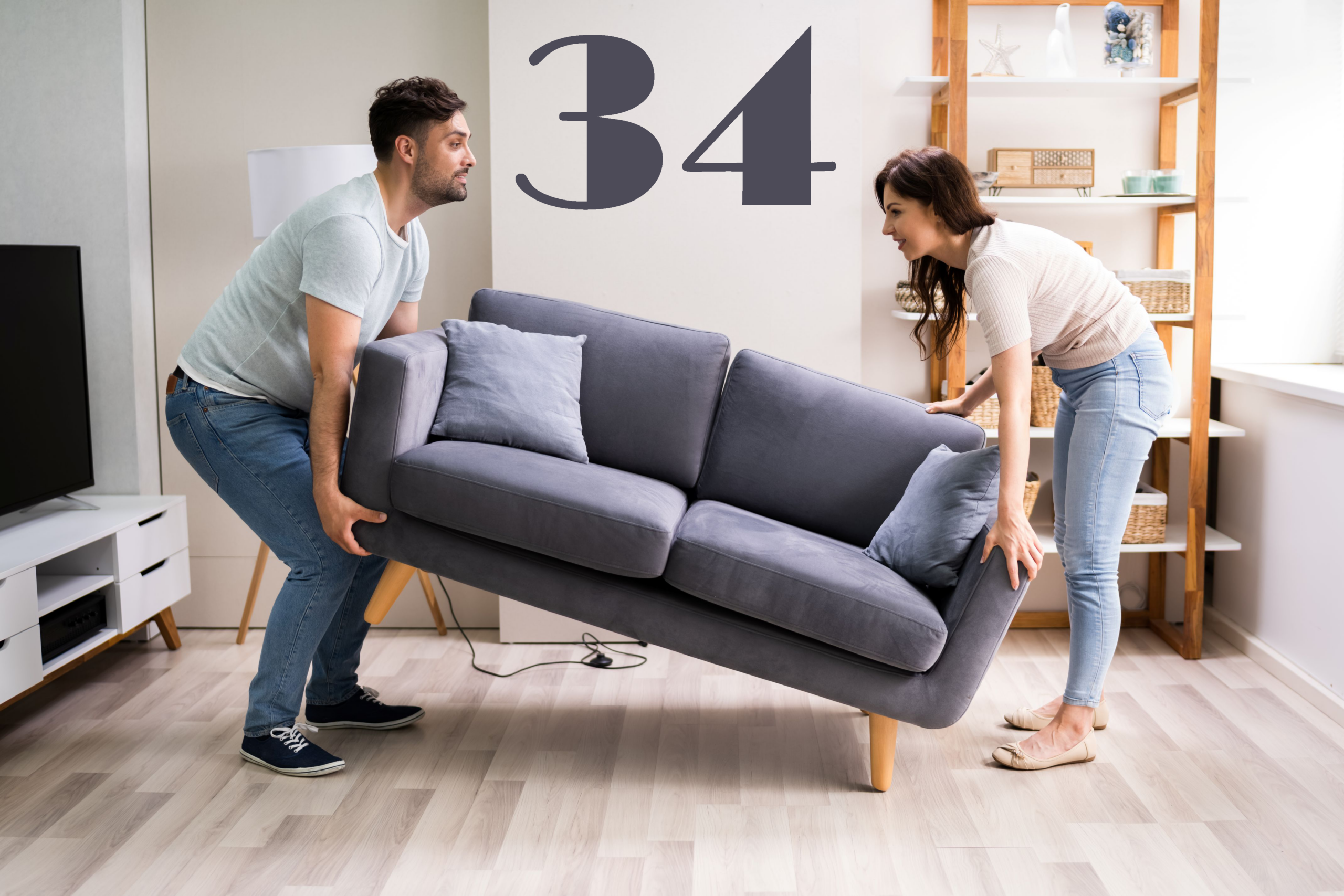 Man en vrouw tillen samen een grijze bank in een lichte woonkamer, met groot cijfer 34 op de muur boven de bank.