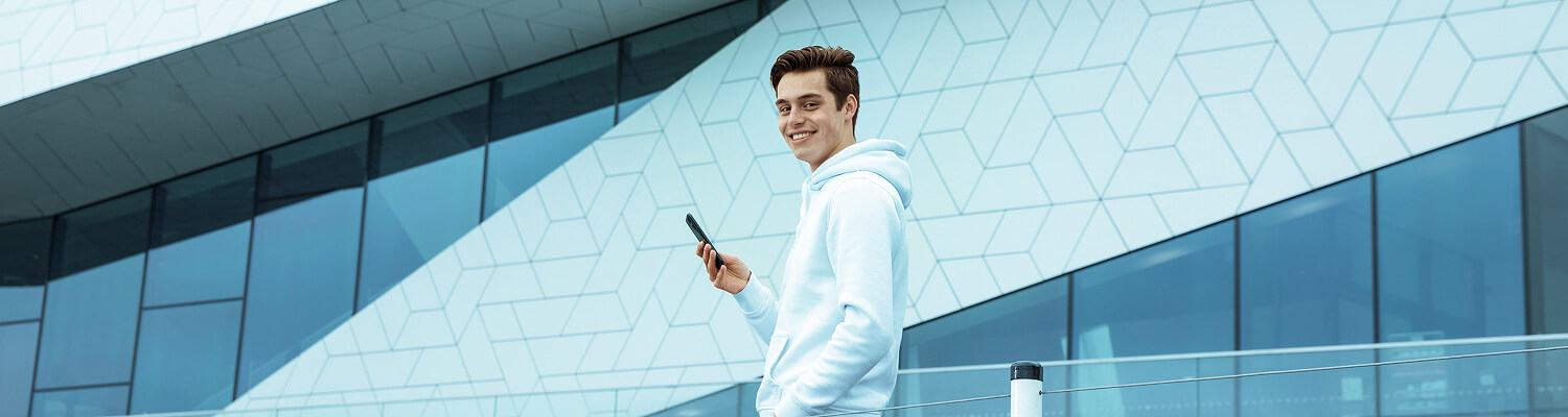 Jonge man in hoodie kijkt glimlachend op terwijl hij buiten bij een modern gebouw zijn smartphone gebruikt voor mobiel bankieren.
