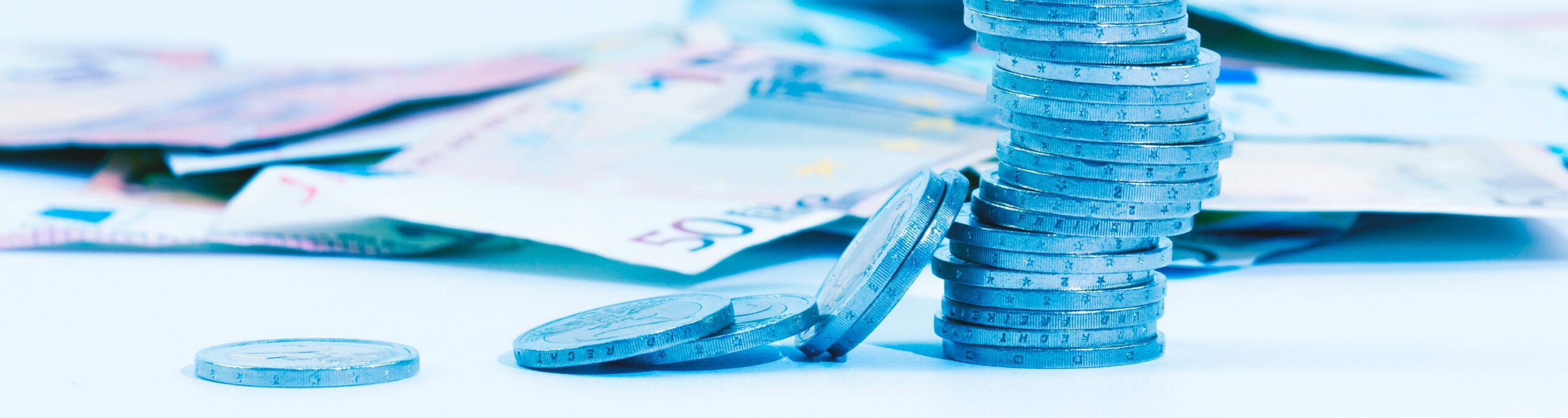 Gestapelde euromunten met losse munten ervoor en eurobankbiljetten op de achtergrond, als beeld van sparen, waardeopbouw en geldstromen.