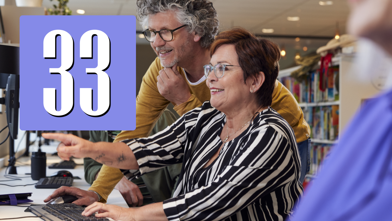 Twee oudere deelnemers werken samen achter een computer in een bibliotheekachtige omgeving, waarbij uitleg wordt gegeven en digitaal gebruik wordt ondersteund.