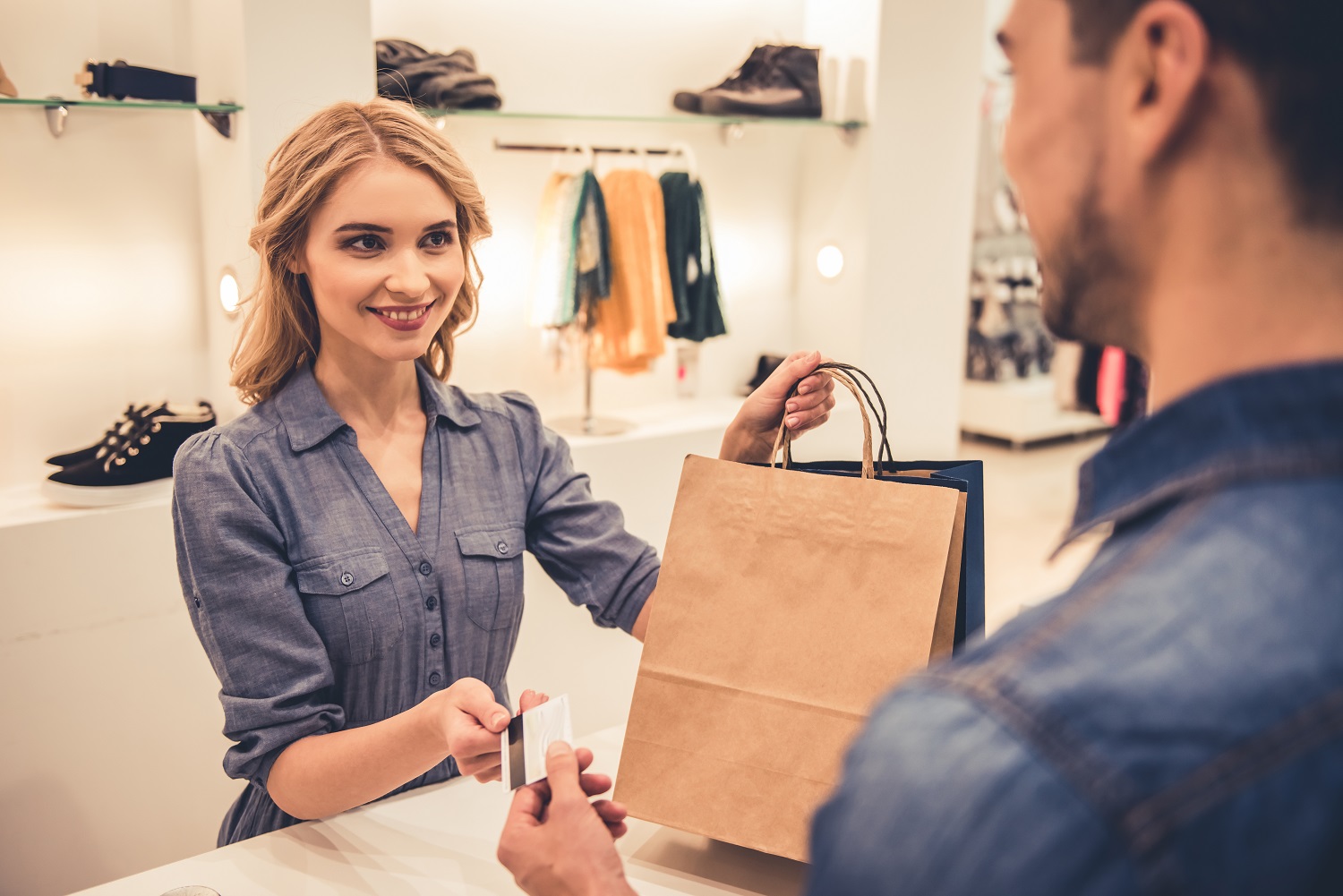 Winkelmedewerker geeft klant een papieren tas aan de kassa terwijl hij een betaalpas overhandigt in een lichte modewinkel.