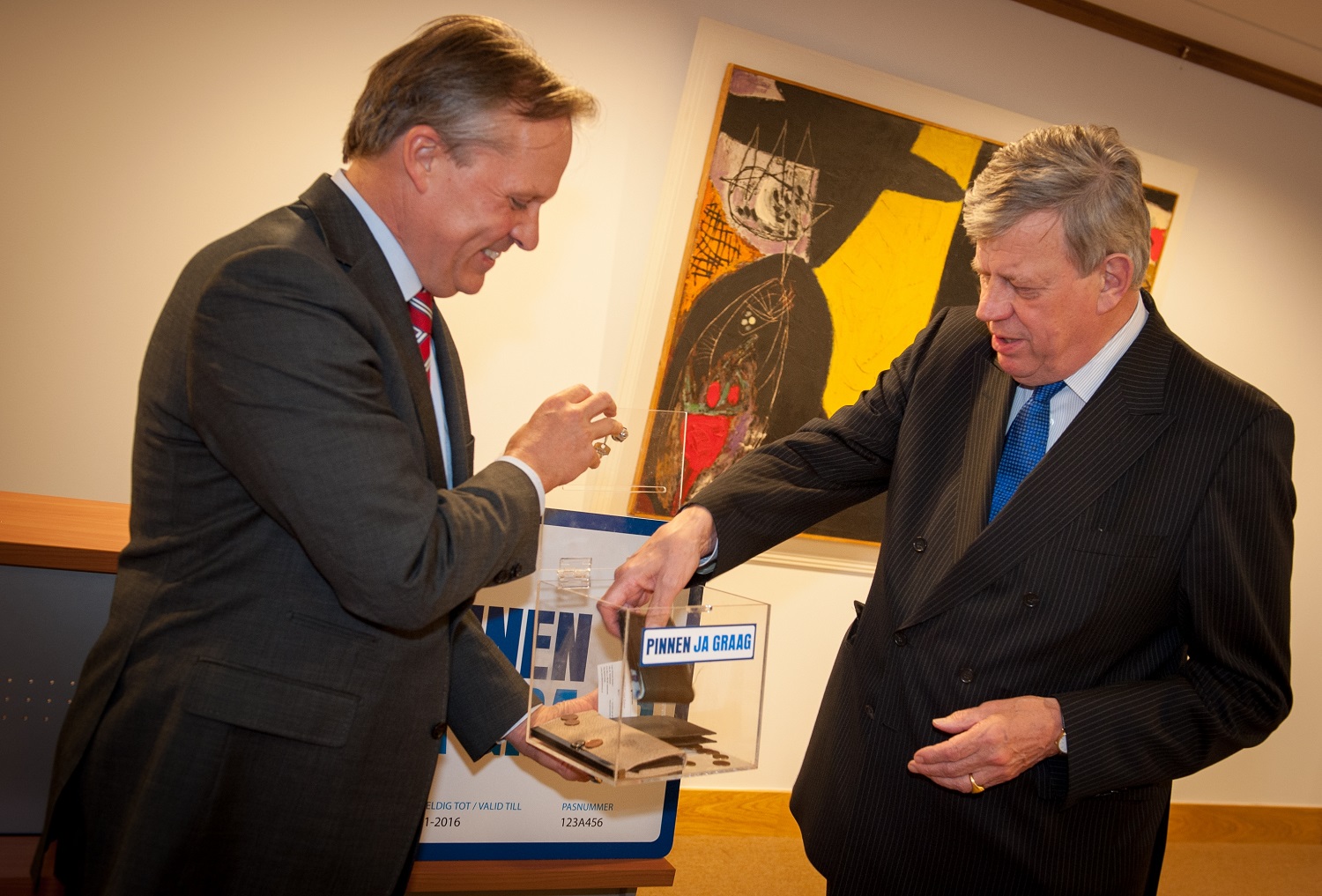 Twee mannen in pak stoppen contant geld in een transparante inzamelbox met het opschrift “Pinnen ja graag”, tijdens een publieke bijeenkomst over betalen.